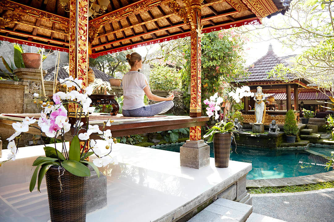 Woman meditating under ornate Balinese pavilion beside peaceful garden pool.
