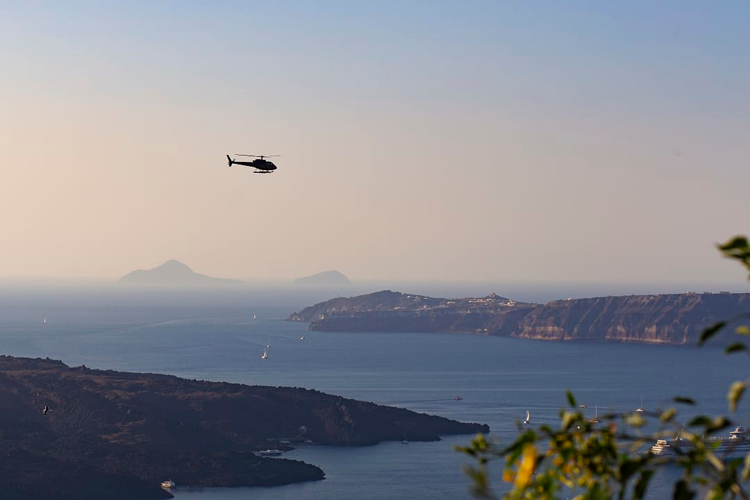 Helicopter flying over deep blue sea near a volcanic coastline.