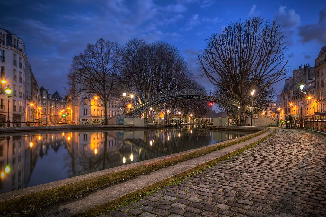 Nighttime scene along the Canal Saint-Martin in Paris during the Christmas season, with lights reflected in the still waters and silhouetted trees lining the cobblestone path