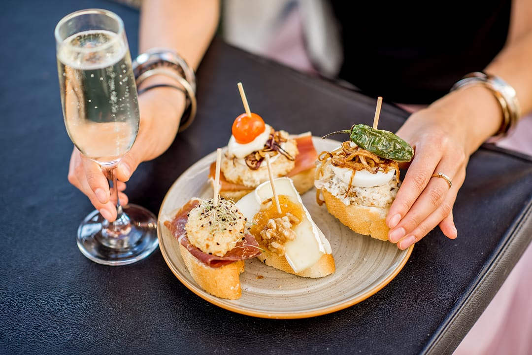 Plate of Spanish pintxos with a glass of cava on an outdoor table, held by a woman