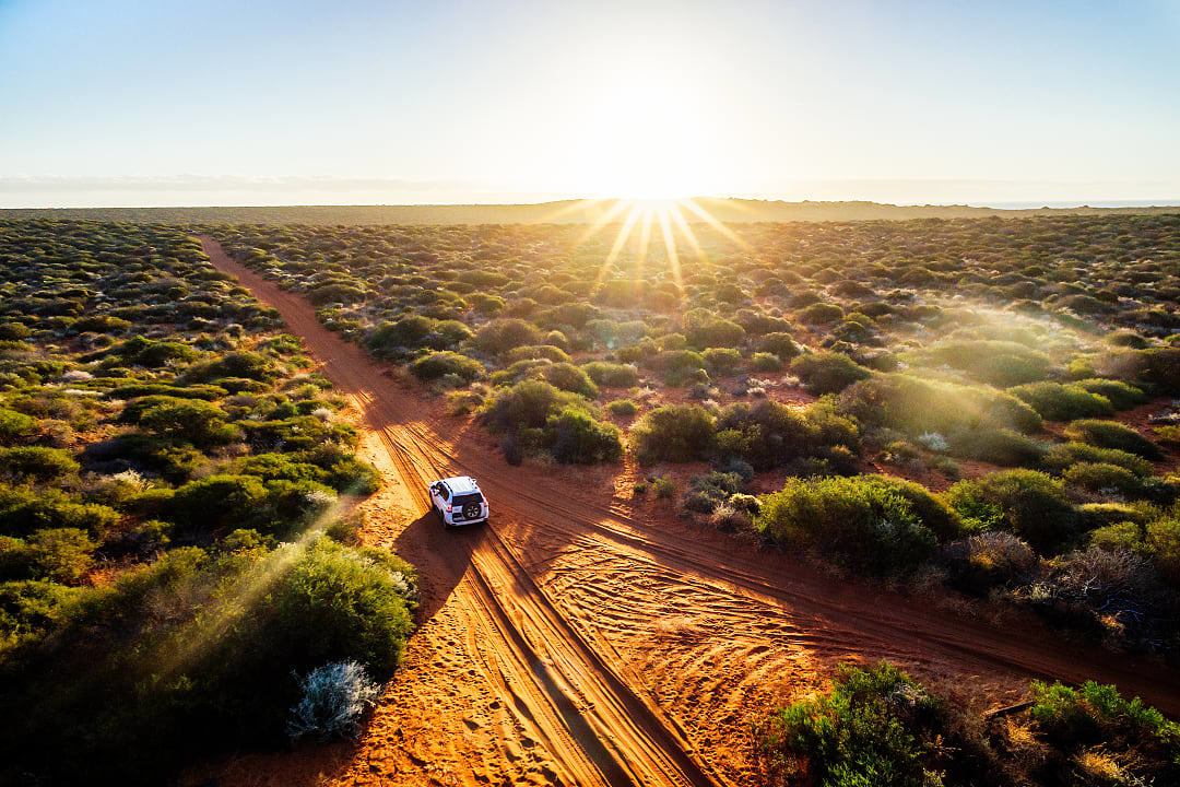 A white luxury 4x4 vehicle driving on a dusty red dirt track through the vast Australian Outback at sunset.