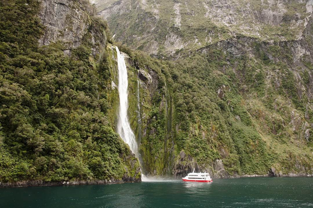 Tour boat approaches towering waterfall cascading down steep green cliffs.