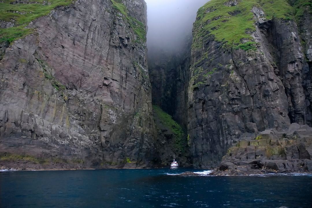 Boat sailing beneath the dramatic Vestmanna Sea Cliffs, with towering rock walls and mist above, Faroe Islands.