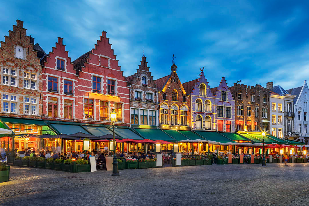 Row of colorful gabled buildings with illuminated outdoor restaurants at dusk.