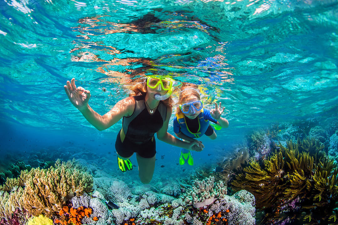Mother and child snorkeling in clear blue water above vibrant coral reefs and marine life.