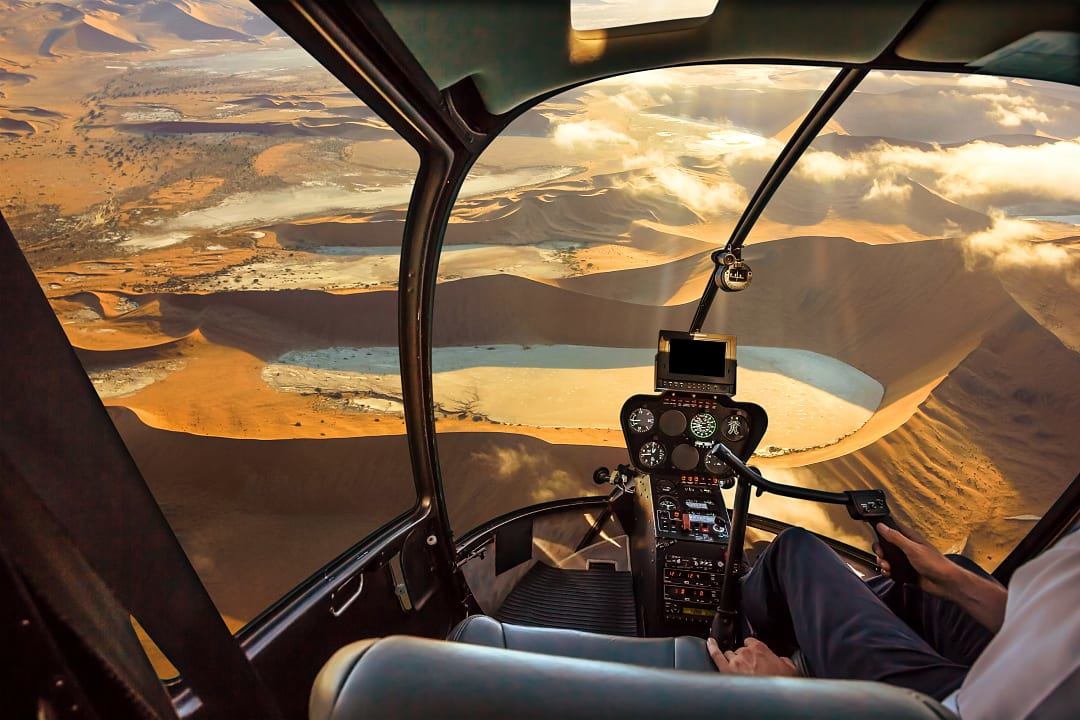 A pilot flying a helicopter over vast red sand dunes, offering a panoramic view of the dramatic desert landscape below.