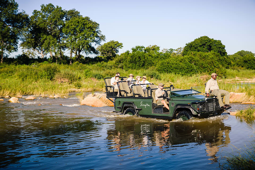 Safari vehicle crossing a shallow river with guides and travelers observing wildlife in a lush, sunlit landscape.