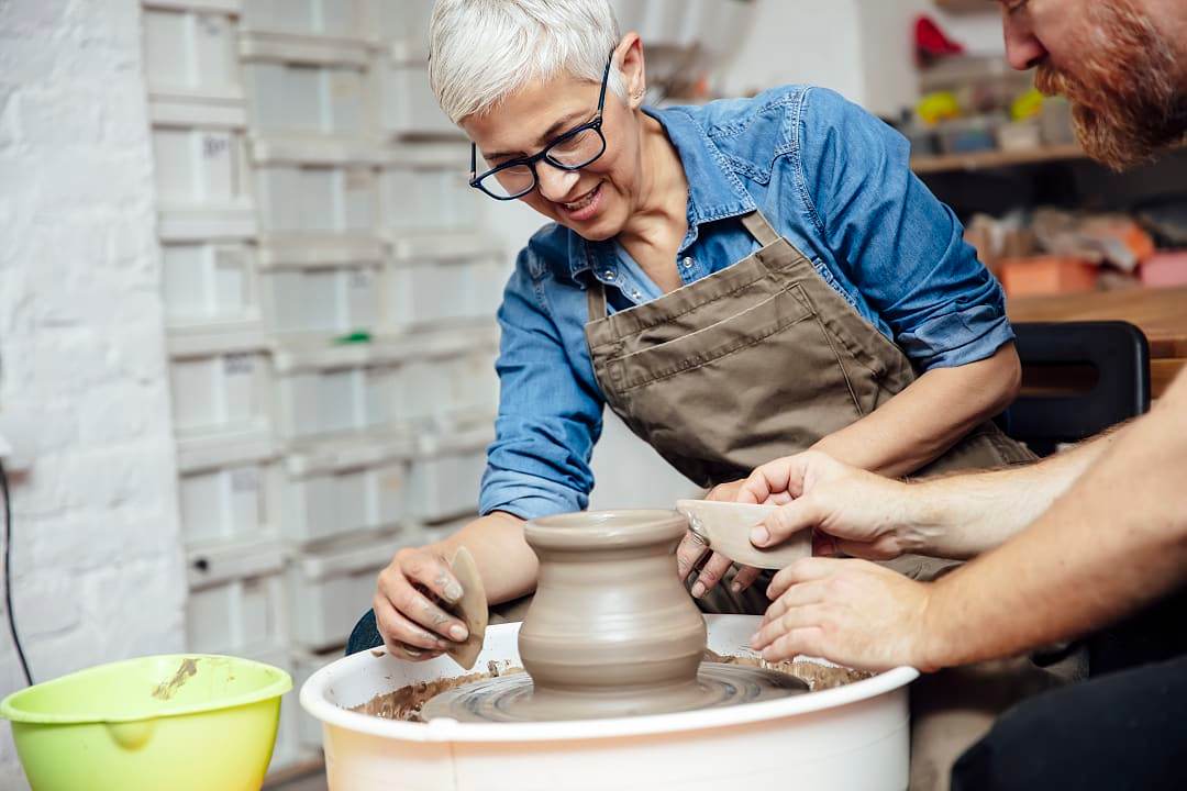 A senior woman with glasses smiling while working with a pottery wheel alongside an instructor in a ceramics workshop.