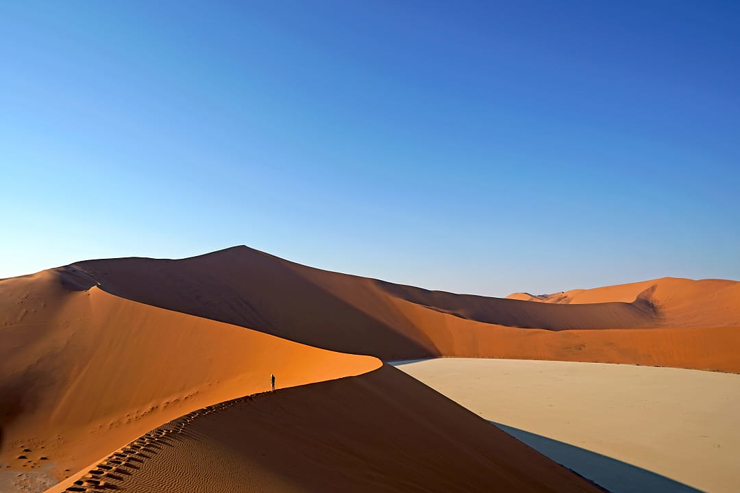 A person walking along the crest of a towering red sand dune under a clear blue sky, showcasing the vast beauty and scale of the desert landscape.