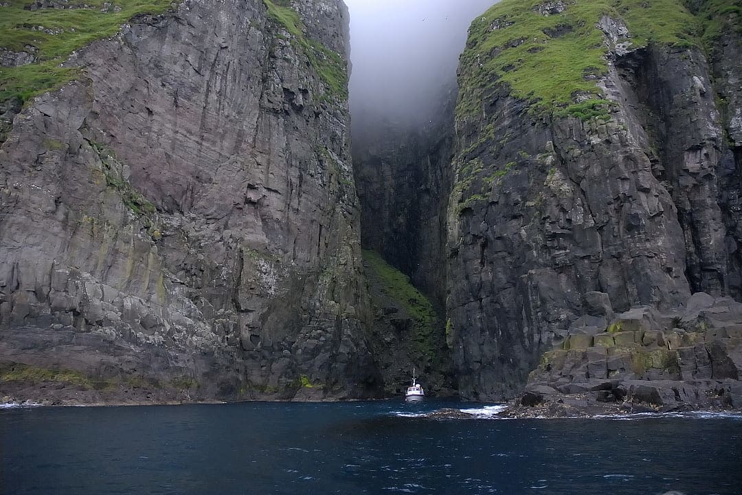 Boat sailing beneath the dramatic Vestmanna Sea Cliffs, with towering rock walls and mist above, Faroe Islands.