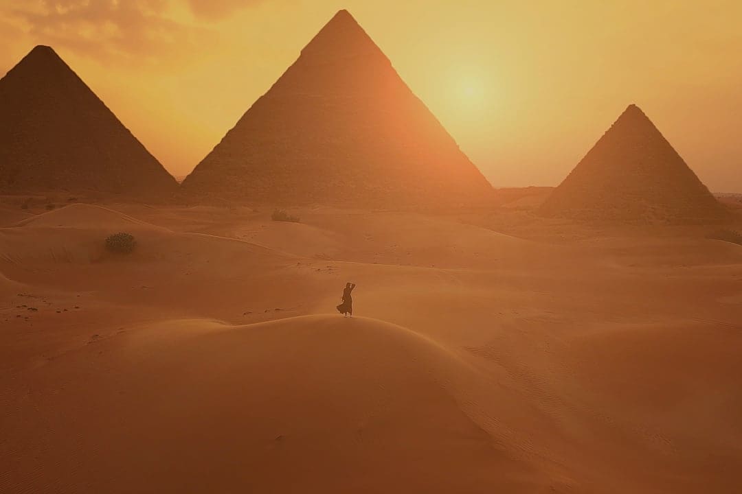 Traveler walking through the desert at sunset with the pyramids of Giza silhouetted in the golden light.