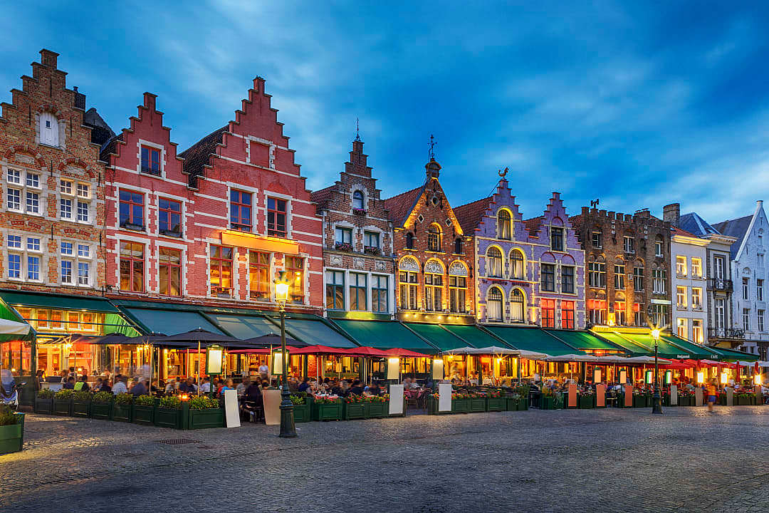 Row of colorful gabled buildings with illuminated outdoor restaurants at dusk.