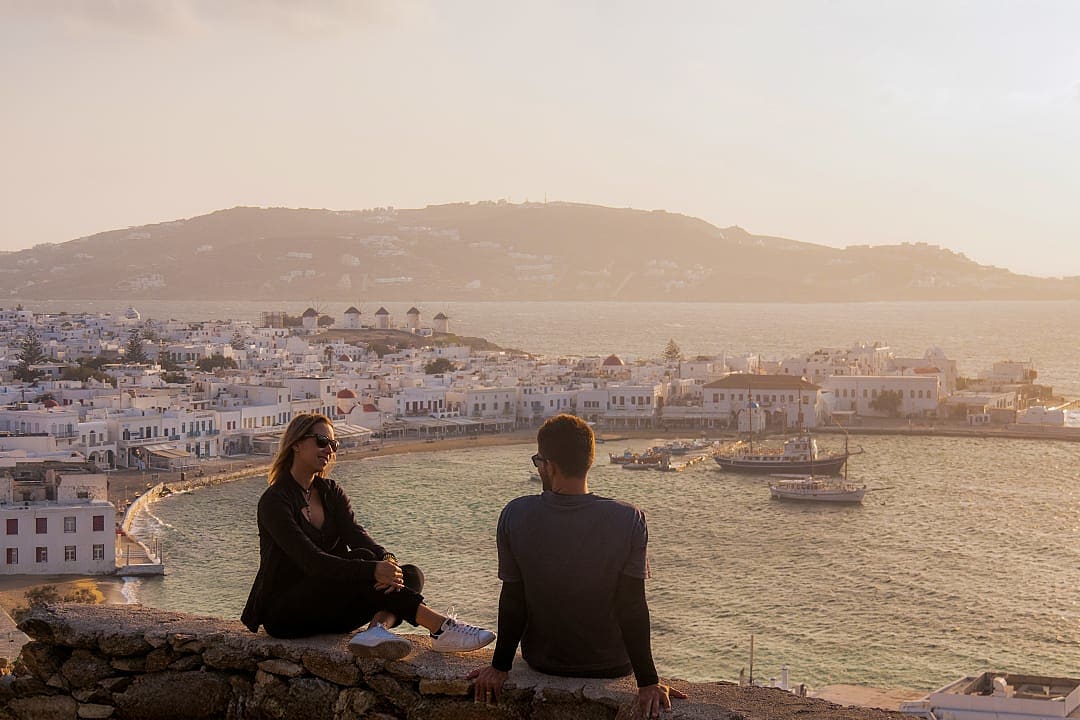Couple enjoying sunset over Mykonos harbor and whitewashed townscape.