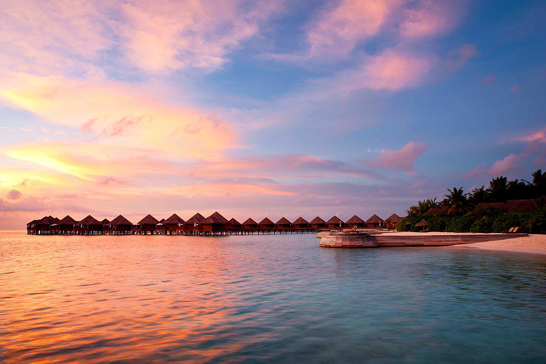 Sun setting over overwater bungalows, mirrored in the crystal-clear waters of the Indian Ocean.