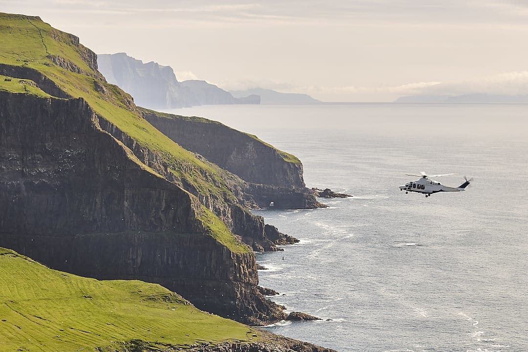 Helicopter flying above the rugged coastal cliffs of Vágar, Faroe Islands, overlooking the North Atlantic Ocean.