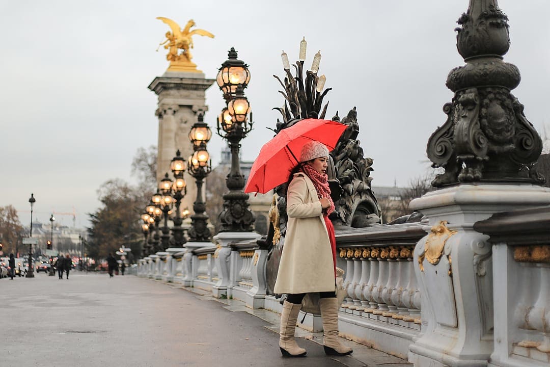 Person with red umbrella walking on Pont Alexandre III in Paris, with ornate lamps and statues on a cloudy day during the Christmas season.