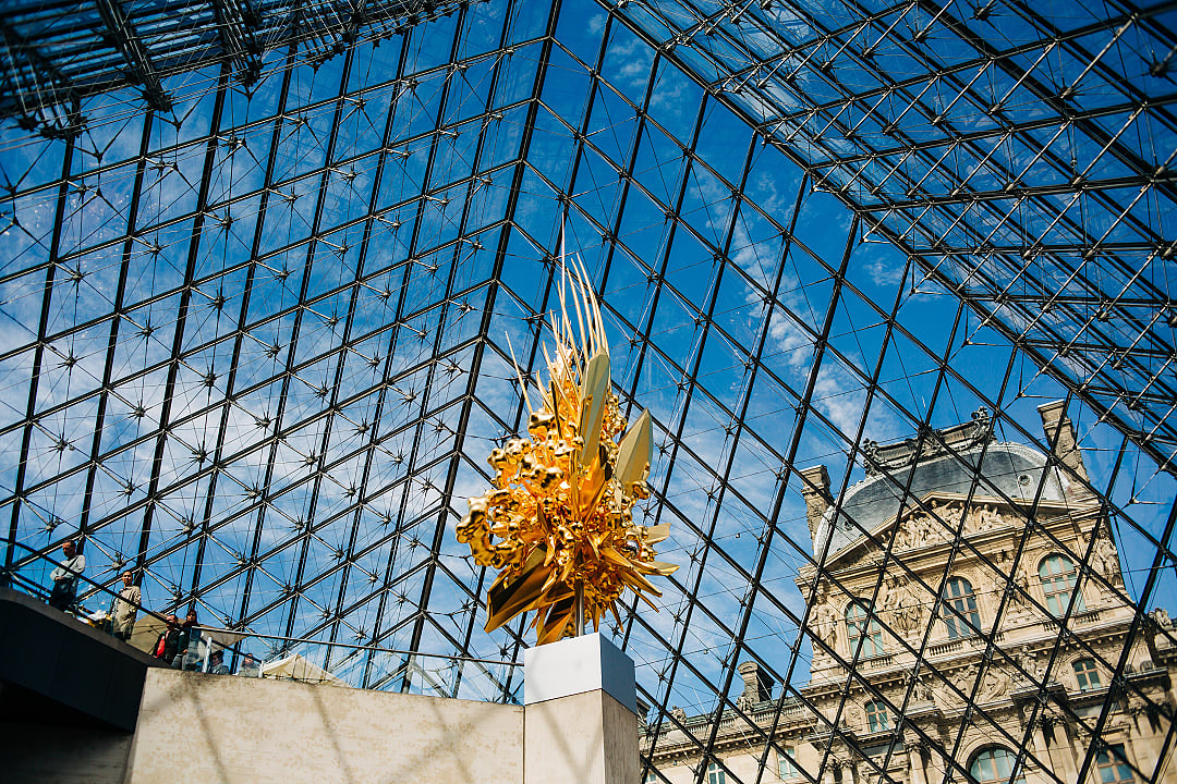 The Louvre’s glass pyramid reflects sunlight over a golden artistic sculpture.
