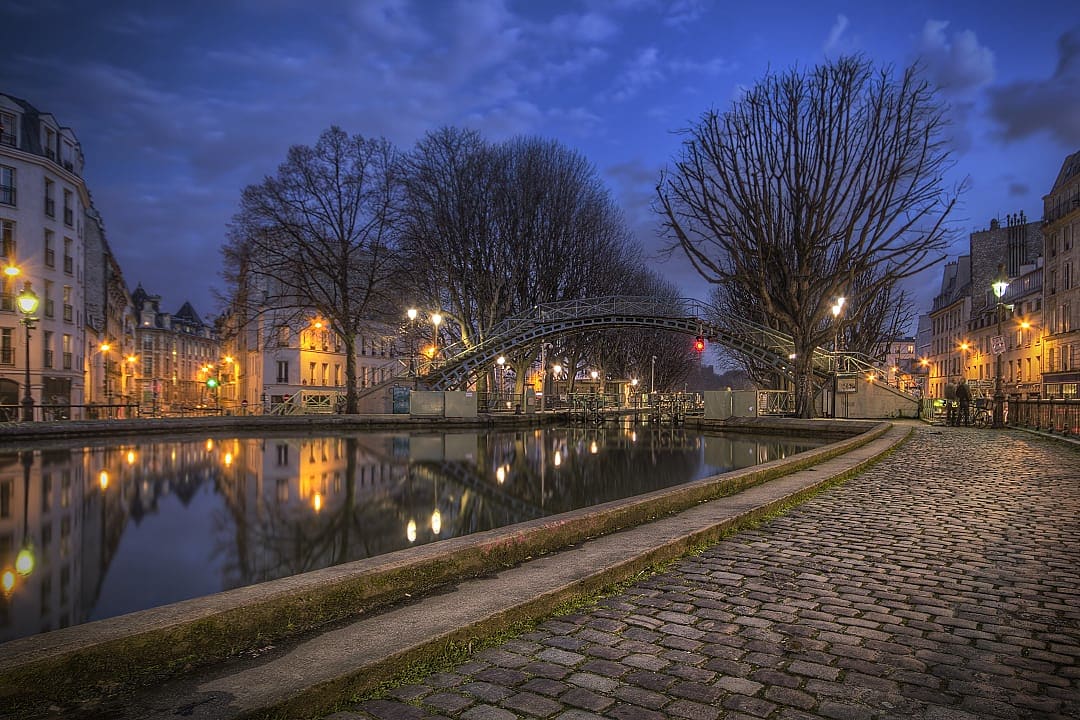 Nighttime scene along the Canal Saint-Martin in Paris during the Christmas season, with lights reflected in the still waters and silhouetted trees lining the cobblestone path
