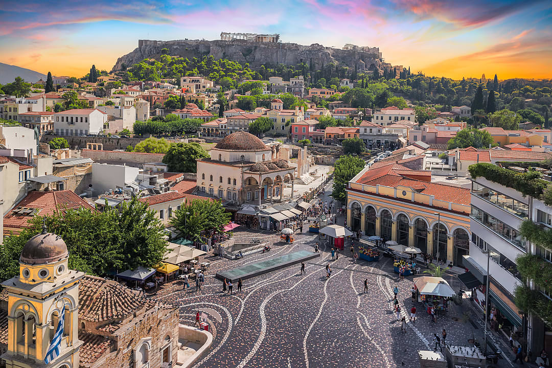 Bustling square beneath Acropolis, vibrant rooftops, historic charm, sunset glow.