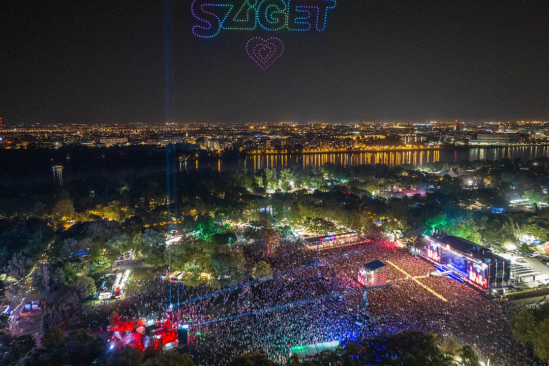 Aerial view of Sziget Festival at night with colorful lights, crowds, and a vibrant cityscape backdrop