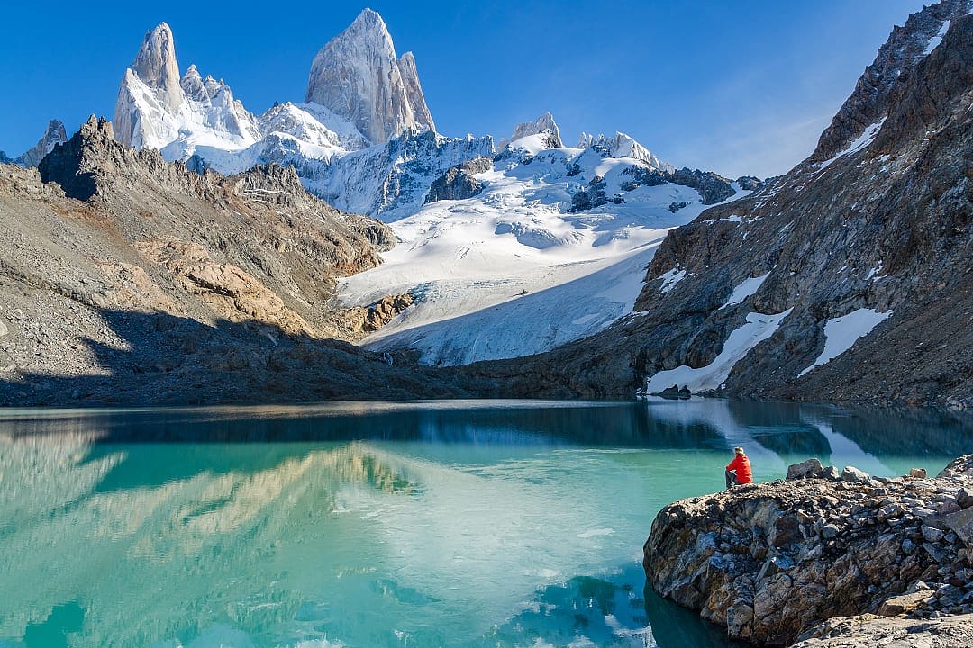 A man hiking on Mount Fitz Roy, looking at the reflective turquoise waters and mountain peaks.