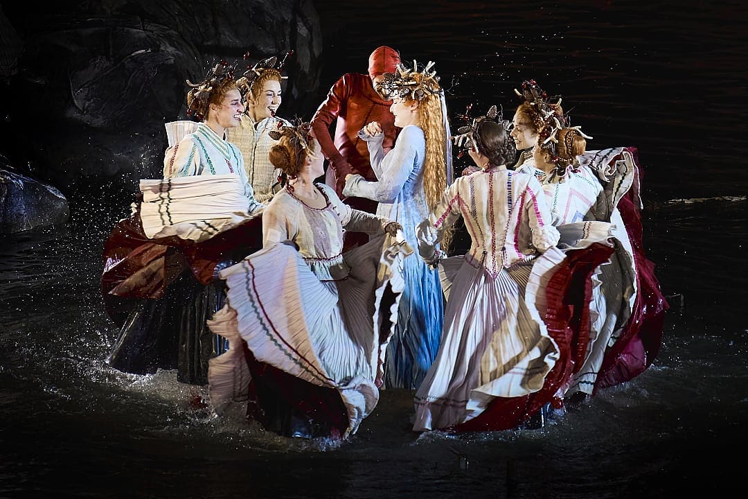 Performers in colorful, ornate costumes stand on a dark stage, creating a visually striking scene