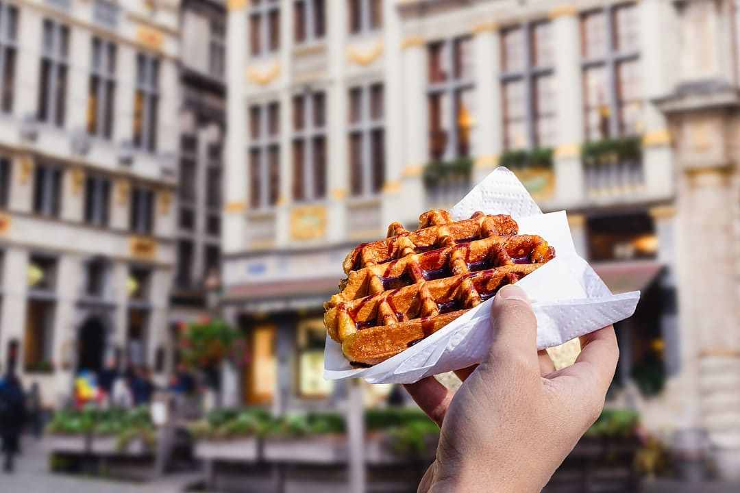 Hand holding a pearl-sugar-studded Liège waffle in a city square.