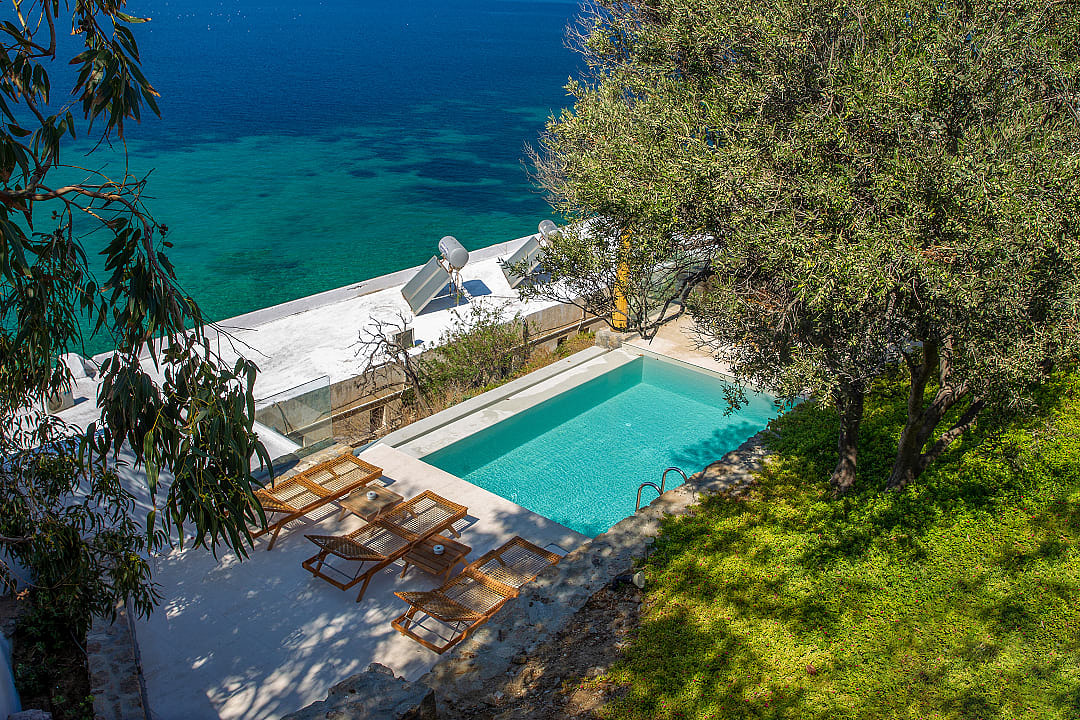 Luxury villa pool and loungers beside clear turquoise Greek sea.