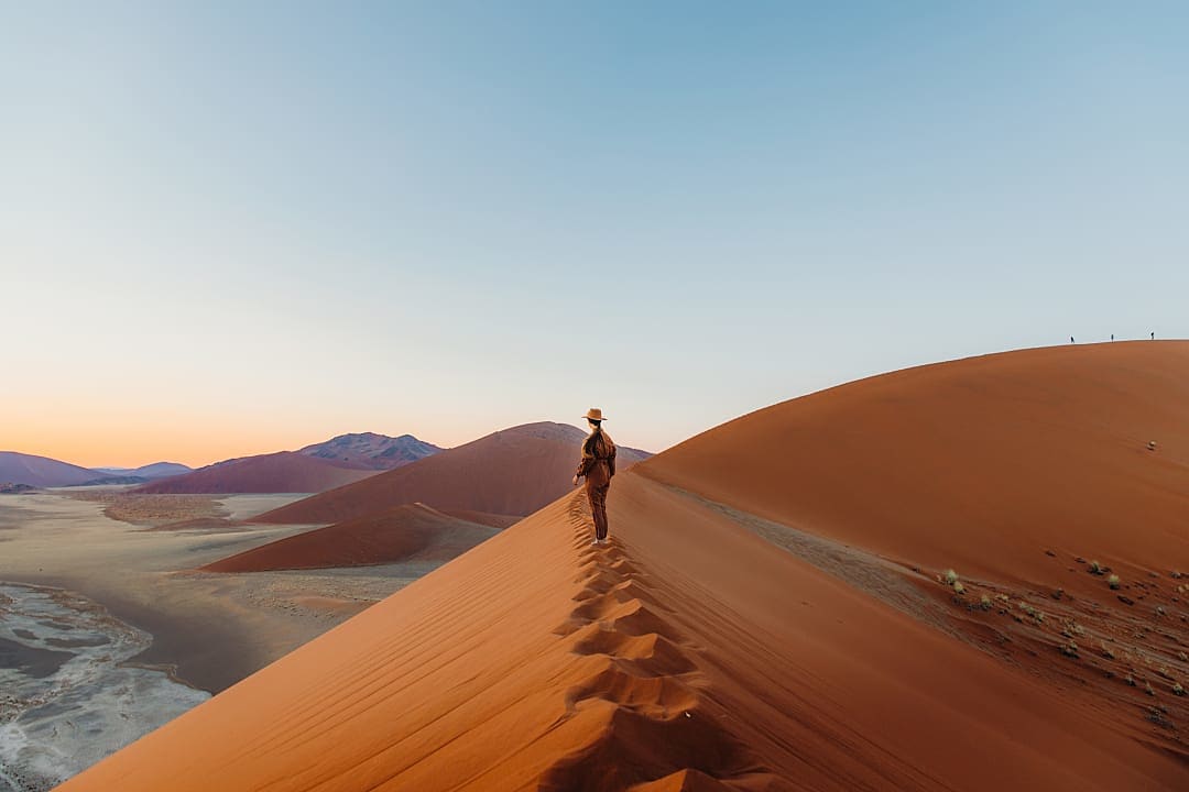 Hiker walks along towering red dunes in Namib Desert sunrise.