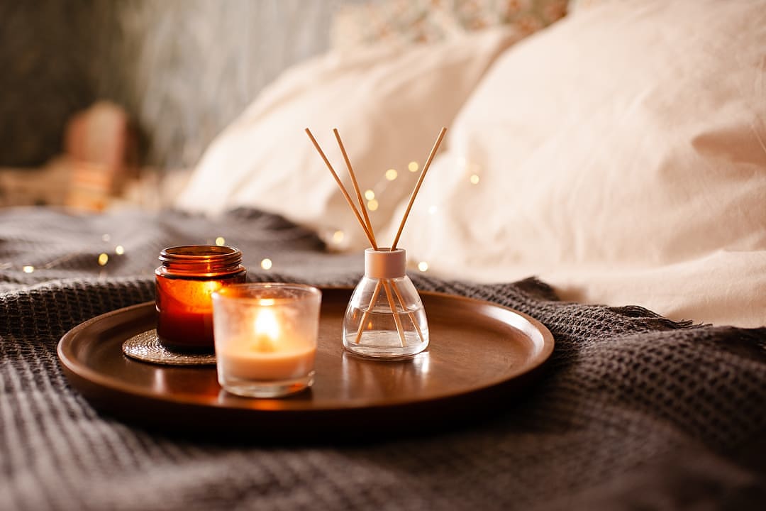 Candles and an aroma diffuser arranged on a wooden tray atop a soft blanket in a cozy, softly lit bedroom.