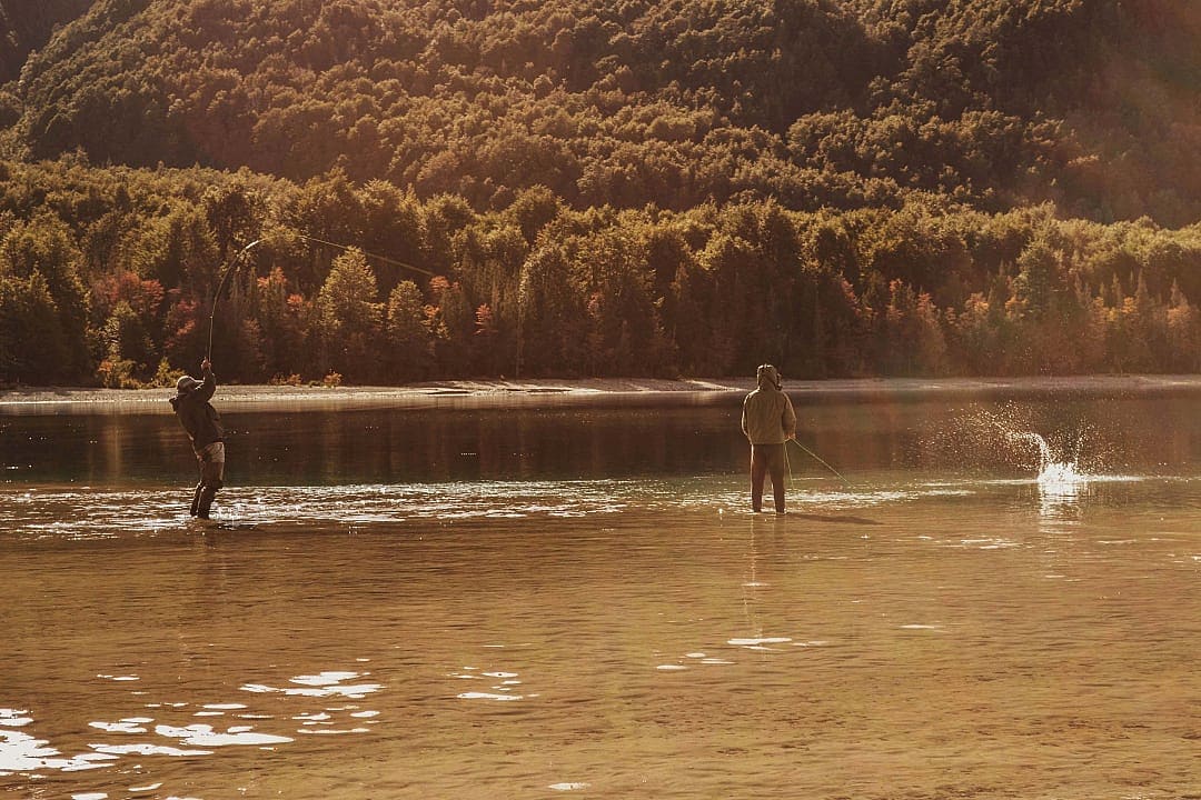 Two anglers fly fish in a peaceful lake surrounded by forest.