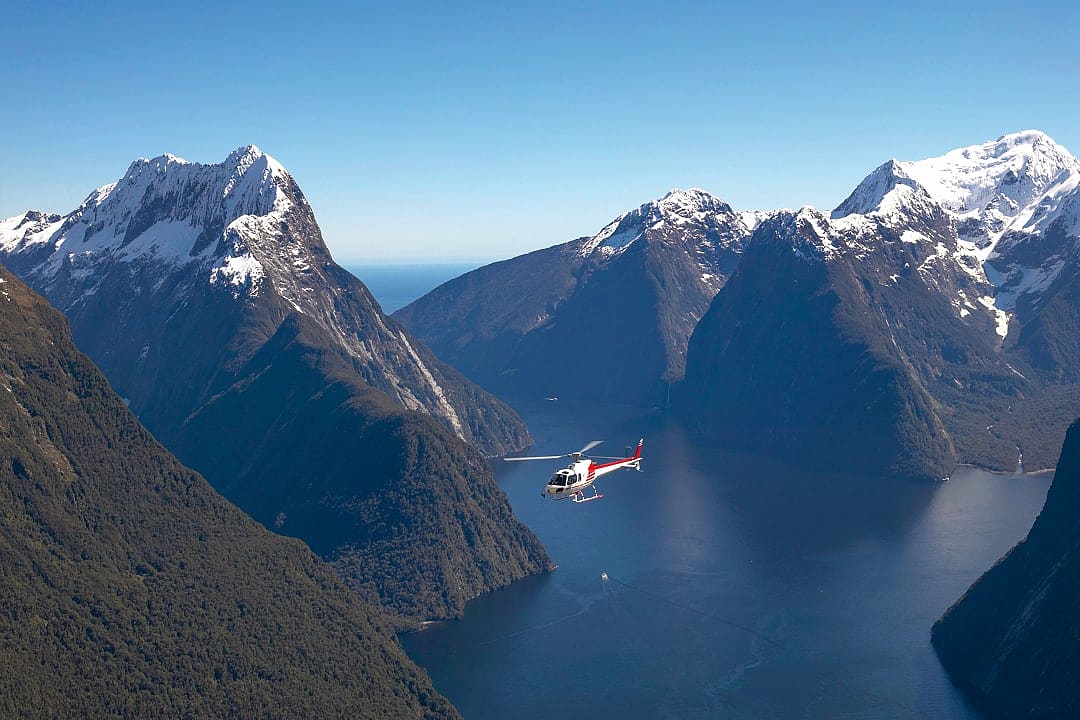 Helicopter soars above Milford Sound’s dramatic fjord and snowy peaks.