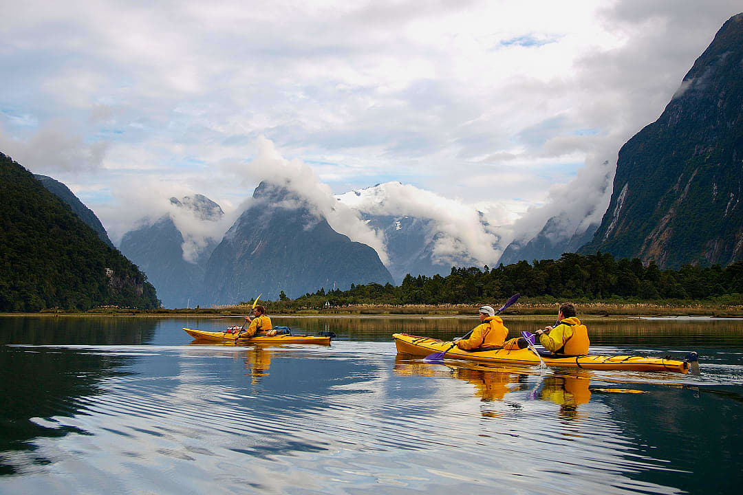 Kayakers explore misty fjords beneath dramatic peaks and drifting clouds.