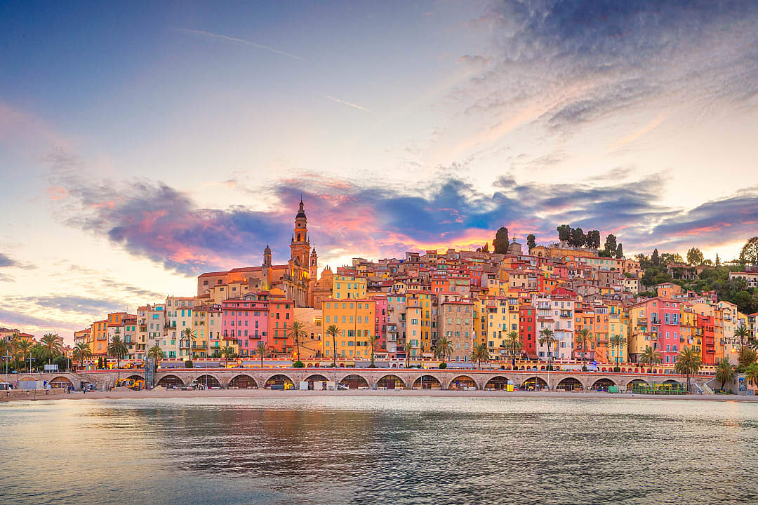 Scenic view of Menton, France, with colorful Mediterranean-style buildings along the coast; palm trees and sandy beach in the foreground, with a backdrop of clear blue skies and lush hills