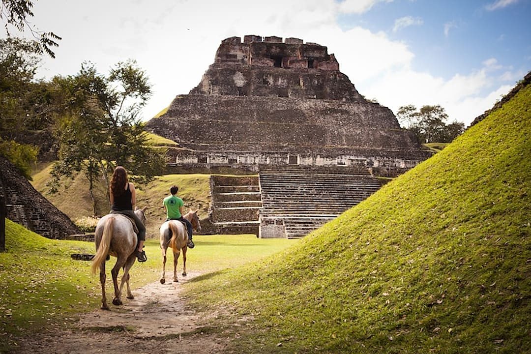 Two people horseback riding towards the ancient Mayan ruins of Xunantunich in Belize on a sunny day