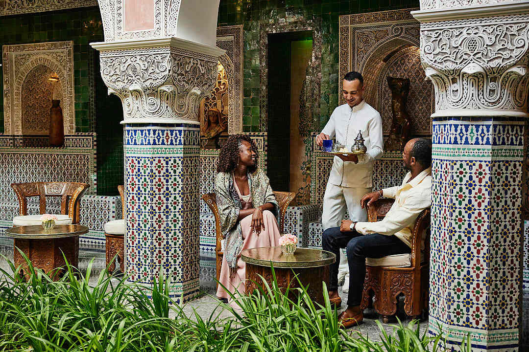 A couple sits in an ornate, tiled courtyard of a traditional Moroccan riad while a staff member serves them.