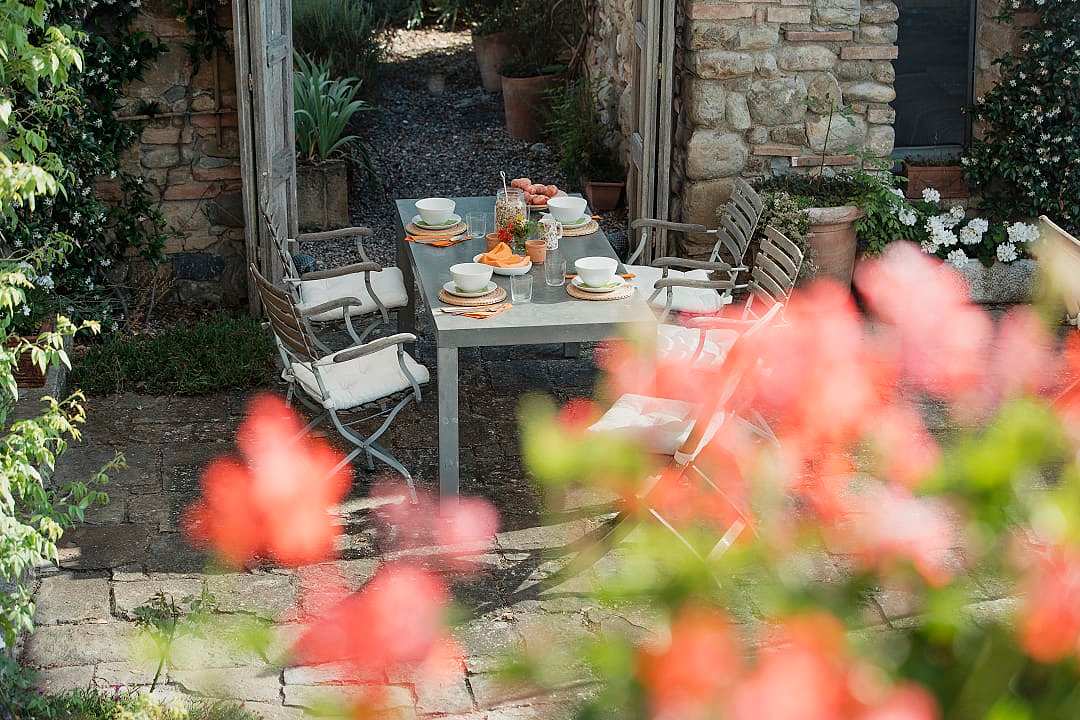 A wooden garden table set for breakfast with coffee, pastries, and fruit in a sunny courtyard.