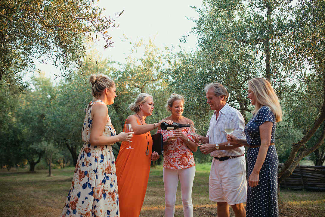 A group of friends stands in a sunny Italian olive grove or vineyard, smiling and talking while holding glasses of wine.