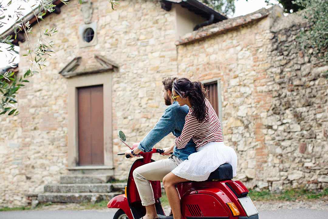 Couple riding red scooter past rustic stone buildings in rural Italy.