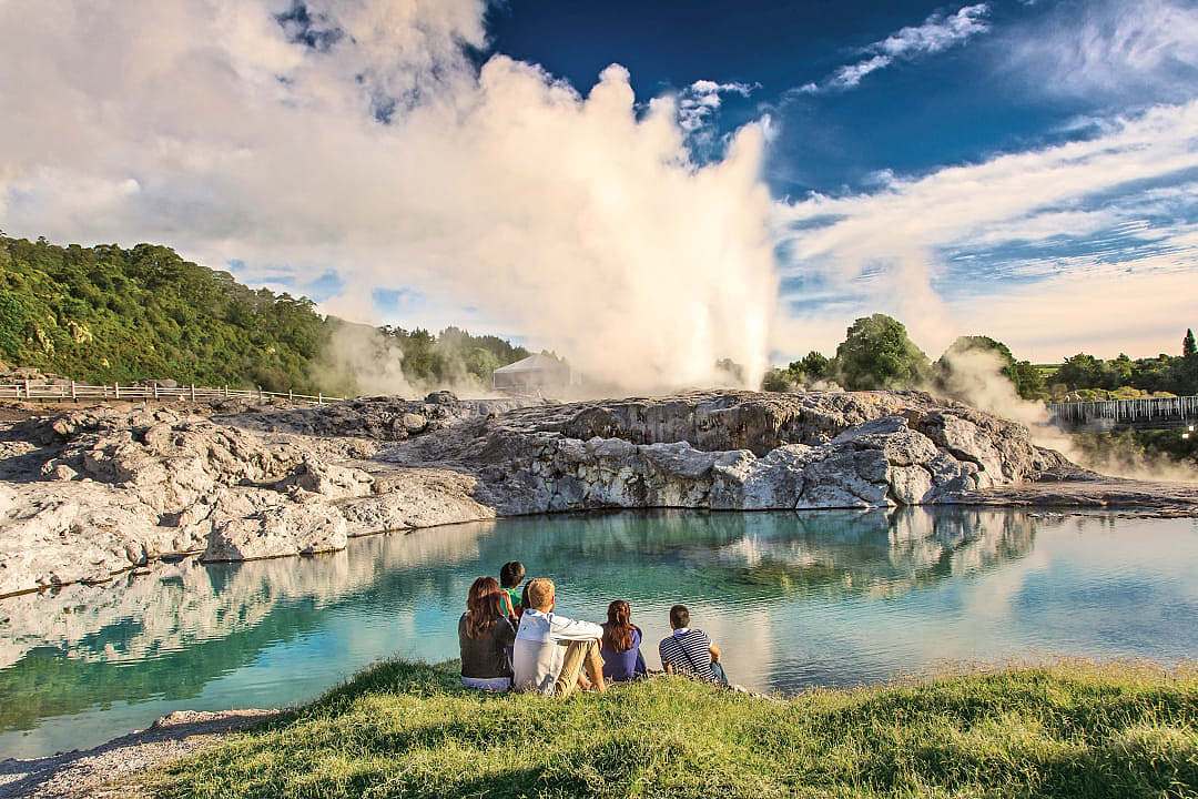 Tourists watching the Pohutu Geyser erupt at Te Puia geothermal park in Rotorua, New Zealand, with steam rising from rock formations and a clear blue pool in the foreground against a lush, green landscape and blue sky
