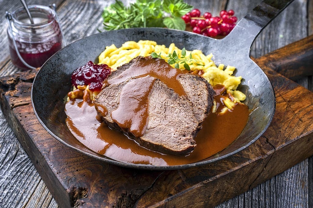 Traditional braised marinated German beef sauerbraten with spaetzle and lingonberry jam in a spicy brown sauce as a closeup in a rustic cast iron frying pan