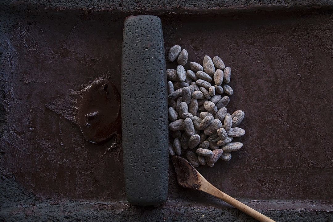 Traditional Mayan cacao grinding with raw cacao beans, stone grinder, and wooden spoon on a dark surface