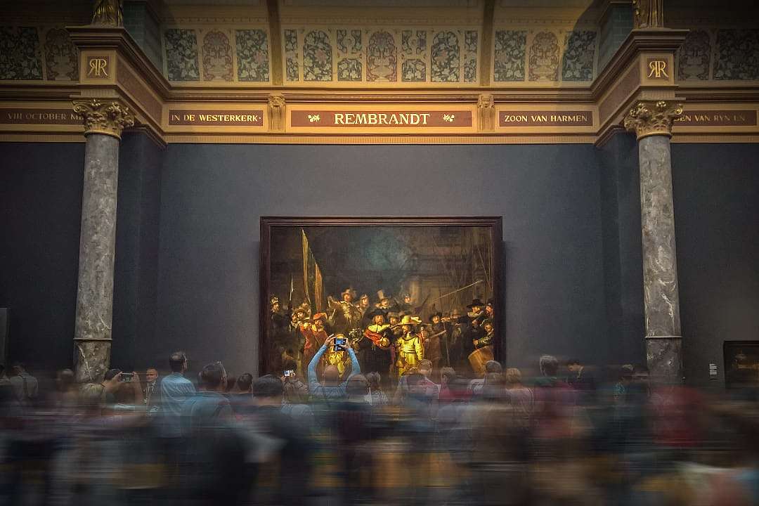 Crowds gathered in front of Rembrandt's large-scale masterpiece, The Night Watch, in a grand gallery.
