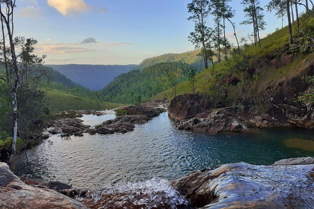 Scenic mountain river with clear water, surrounded by lush green hills and trees under a vibrant sky.
