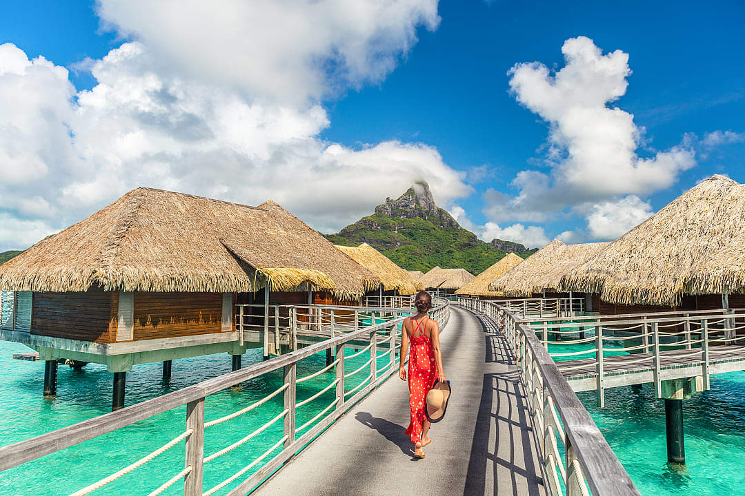 A woman strolling along a wooden boardwalk between overwater bungalows at a turquoise lagoon resort in Bora Bora.