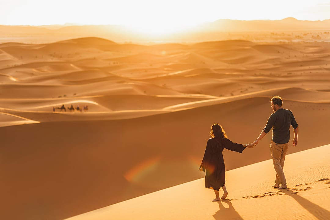 Couple walking hand in hand across Sahara sand dunes at sunset in Morocco.