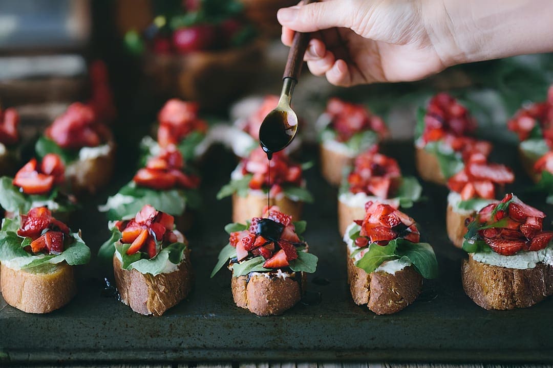 Hand drizzling balsamic glaze over crostini with ricotta, arugula, and fresh strawberries