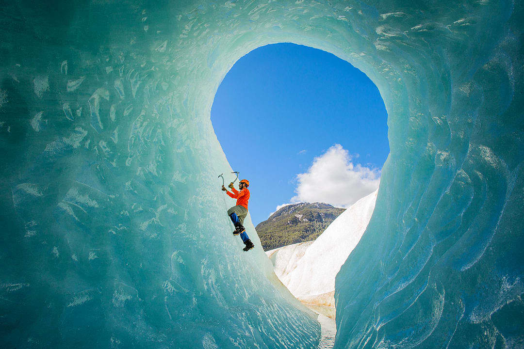 Climber scaling an ice cave wall in Patagonia with a clear blue sky and mountains in the background