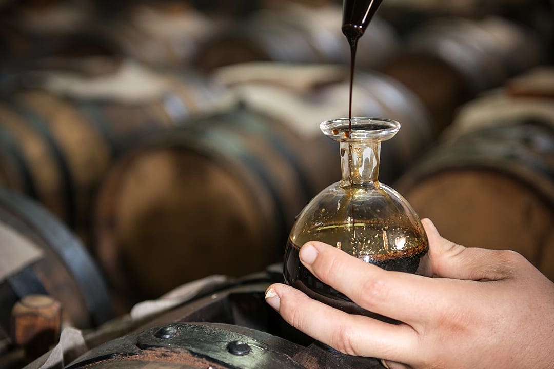 Traditional balsamic vinegar being poured into a glass bottle, aged in wooden barrels in the background
