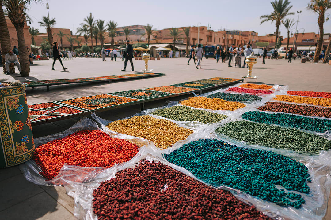 Colorful spices displayed in an open-air market with people walking in the background.